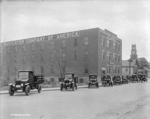 View across street towards a fleet of delivery trucks parked at an angle in a long line in front of an International Harvester of America building. There are men sitting inside the trucks.