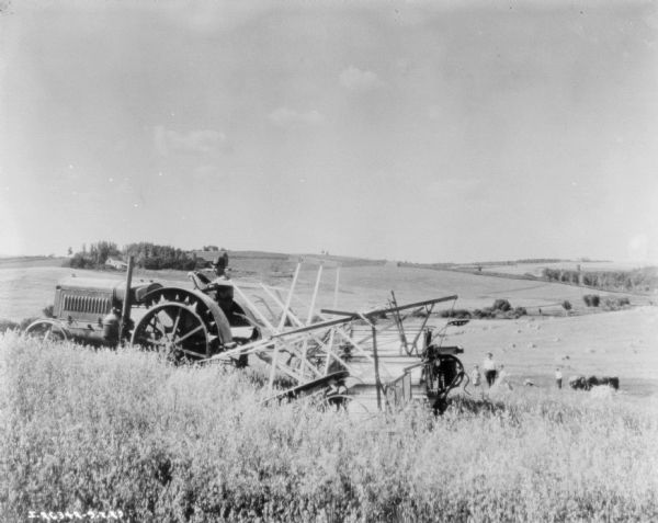Left side view of a man using a tractor-drawn binder in a field. In the background down a slop a man and two children are watching. Harvested grain is in the field below. Farm buildings are among trees on a hill in the background.