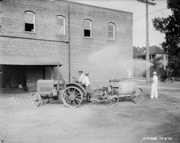 View across yard towards a man driving a tractor pulling an industrial paint sprayer. Two men are standing behind the sprayer holding two hoses with the nozzles high up spraying into the air near a brick building. In the background is an automobile in the street and another large building.