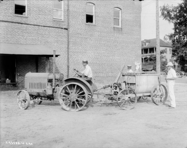 View across yard towards a man driving a tractor pulling an industrial paint sprayer. Two men are standing behind the sprayer holding two hoses with nozzles near a brick building. In the background is an automobile in the street and another large building.