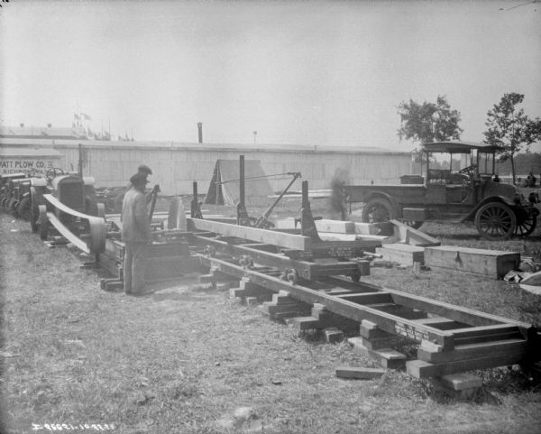 Two men are working with a large saw powered by a tractor on a construction site. There is an automobile on the right, and a tent in the background near a long building. A sign near the building on the far left reads in part: "...ATT PLOW Co. Inc., Richmond, VA." 
