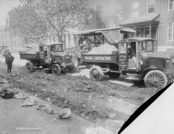 Men with fleet of delivery trucks at a construction site on a street. The trucks on the left and the right have signs painted on the side reading: "Paving Contractors, Carlon Con'st Co., Maplewood, MO." Buildings are in the background.