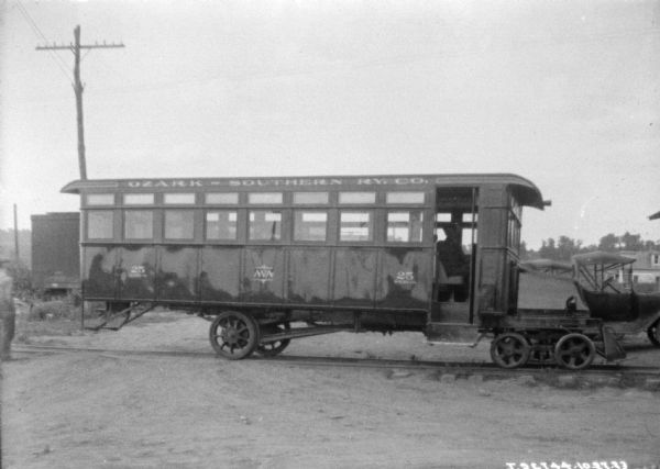 View of a railway car on railroad tracks. Automobiles are parked behind it on the far right. There is a man in the driver's seat of the railway car, and painted along the top side of the car is a sign that reads: Ozark-Southern "Ry. Co." Another man is standing on the far left.