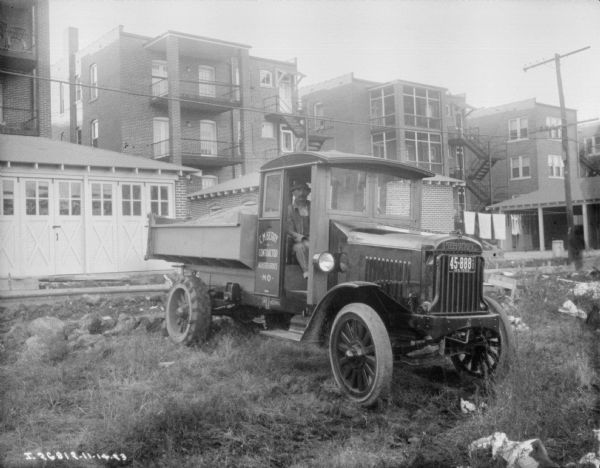 A man is sitting in open doorway of a delivery truck, which has a load of dirt of gravel in the open truck bed. The sign on the passenger side door reads: "C.M. Berry, Contractor, Webster Groves, MO." In the background is a garage, and three-story buildings. 