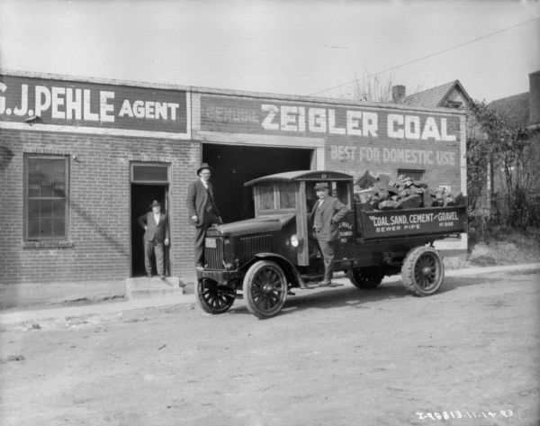 Two men are posing with a coal delivery truck. The sign on the driver;s side door reads, in part: "J. Pehle, ...PLEWOOD, MO." The truck bed if full of material, and a sign on the side reads: "Coal, Sand, Cement and Gravel, Sewer Pipe, WT. 8100." Another man is standing in the doorway of a brick building which has a large, open garage door. "J. Pehle, Agent," is painted on the facade, along with "Genuine Ziegler Coal Best for Domestic Use."