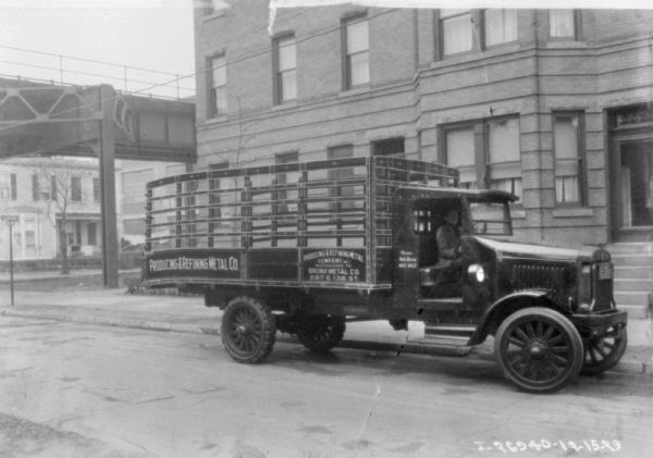 Passenger side view of a man sitting in a delivery truck parked on a street. In the background is an elevated highway or railroad tracks. Buildings and houses are in the background.