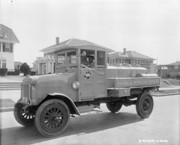 Three-quarter view from front left of a man sitting in a delivery truck parked in front of large homes. The sign painted on the side of the truck reads: "Magnolia Petroleum Company." On the front of the truck is a Texas license plate.