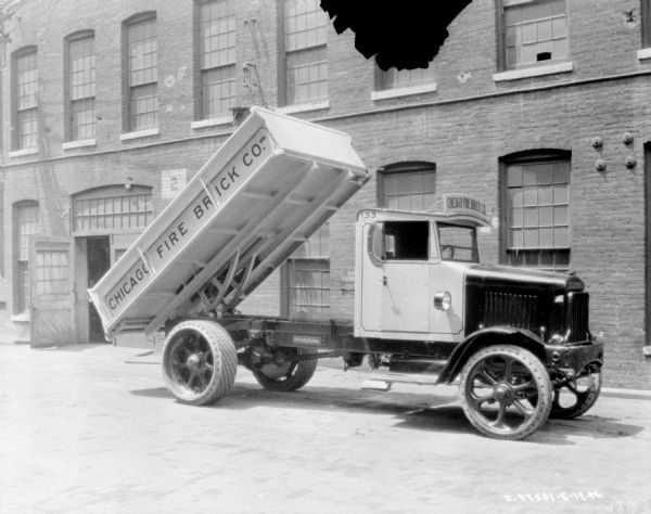 View of a delivery coal truck with the bed raised. A sign painted on the side of the bed reads: "Chicago Fire Brick Co." In the background is a large brick building.