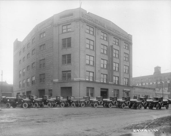 View across yard towards a fleet of delivery trucks parked in front of a large brick building. A sign at the top of the building reads: "International Harvester Co. of America." A one-story building next to it on the right has a sign that reads: "International Motor Trucks." There is a water tank on top of a building in the background.