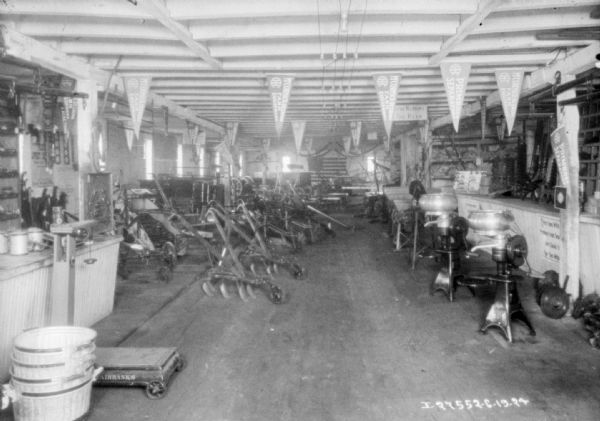 Interior view of a dealership. Agricultural implements are displayed on the floor. On the left is a counter and a floor scale, and on the right are three cream separators lined up near another counter.