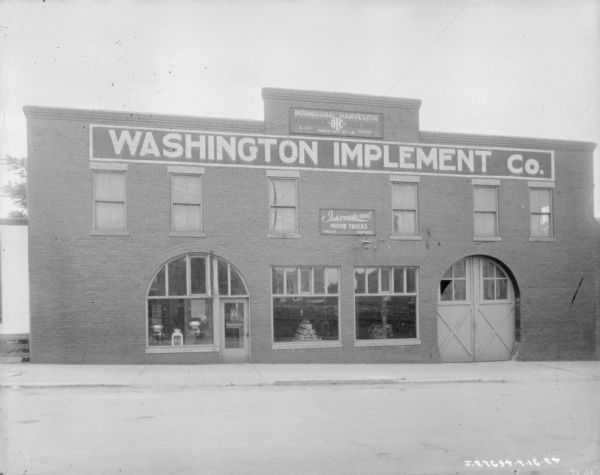 View from across street towards the facade of the Washington Implement Co. dealership. There are cream separators and other equipment on display in the windows.