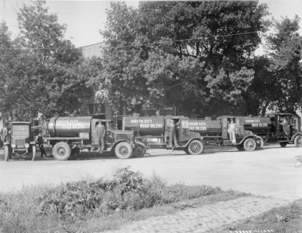View across road towards men posing with oil delivery trucks in front of a large brick building, with a sign in front reading: "International Harvester." The signs painted on the trucks reads: "Gibson City Road Oilers."