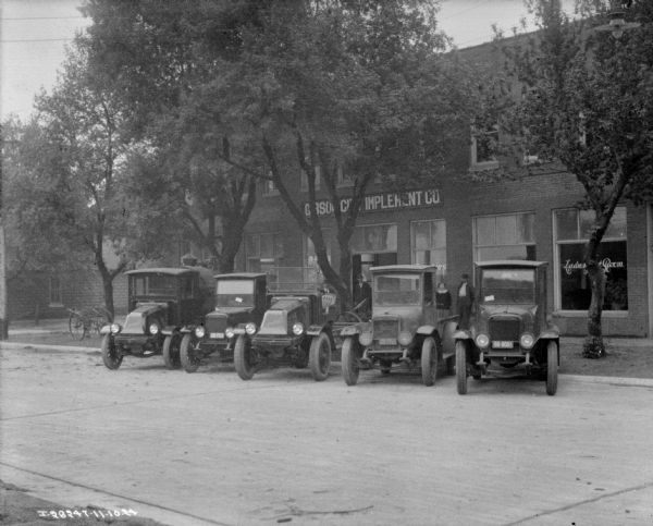 View across street towards delivery trucks parked in front of a dealership. A group of men and women are posing on the sidewalk behind the trucks.