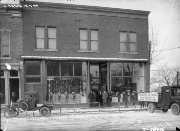 View from across street towards a group of men and women posing in front of a dealership. There are cream separators on display in the show window. The sign on the window reads: "Follmer & Lawrence." A truck on the right has a sign on the side of the truck bed that reads: "McCormick-Deering Primrose Ball-Bearing Cream Separators."