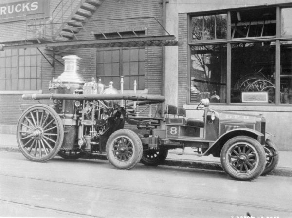 View across street towards a Model SD 1926 fire truck parked along the curb in front of a large building. A sign on the building reads in part: "_RUCK" and a large show window is on the right through which agricultural implements can be seen on display.