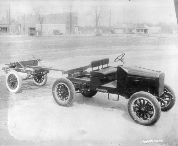 Elevated view of a truck and trailer parked in an open area. There are buildings in the far background. The truck is open, and has a bench seat. There is a small truck bed and an attachment for the trailer which has two wheels.