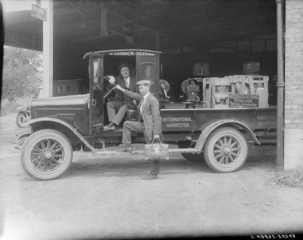 View of a truck sitting in the large, open doorway of a dealership. One of the men is sitting inside the truck with the door open. The other man has one foot on the running board, one hand on the steering wheel, and is holding a briefcase in the other hand. There are cream separators and some crates in the truck bed, along with a sign that reads: "McCormick-Deering Primrose Cream Separator Used On This Farm."