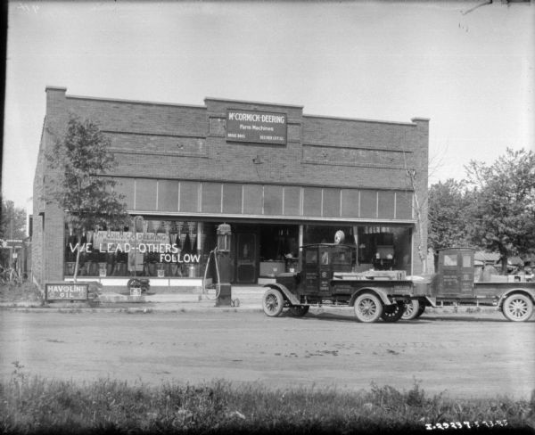 View from across street towards two trucks parked in front of the Hogge Bros. dealership. There are large windows with displays and signs, and a sign painted on the window on the left reads: "We Lead -- Others Follow."