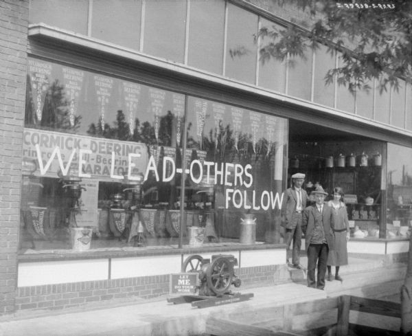 Two men and a woman posed on the steps in front of the Hogge Bros. dealership. There are large windows with displays and signs, and a sign painted on the window on the left reads: "We Lead -- Others Follow."