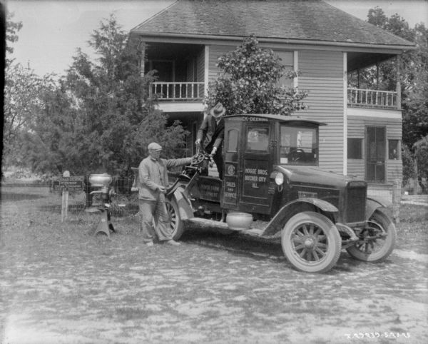 Men moving cream separators using a truck. The sign painted on the passenger side door of the truck reads: "Hogge Bros. Beecher City, Ill." In the background is a house. A man wearing a suit and hat is standing in the open truck bed and pulling an old cream separator up into the truck with the help of another man standing nearby. A new cream separator is sitting on the ground on the left, near a sign posted on the fence which reads: "McCormick Deering Primrose Cream Separator Used On This Farm."