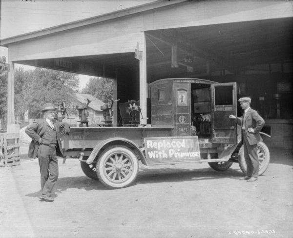 Two men are standing near a service truck with cream separators loaded in the truck bed. A sign on the truck bed reads: "Replaced With Primroses." Behind them is a large, open wood building with what may be a loading dock.
