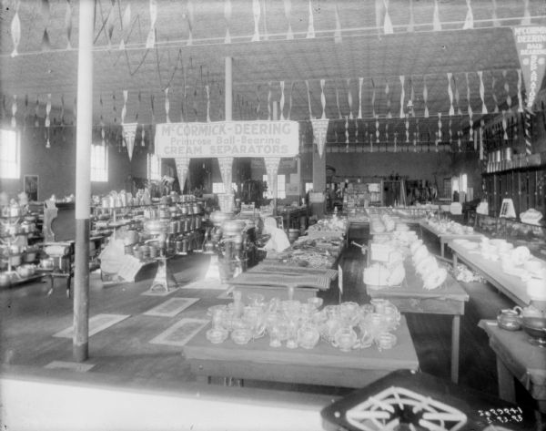 View of merchandise on the floor of a dealership. A large sign hanging from the ceiling reads: "McCormick-Deering Primrose Ball-Bearing Cream Separators." In the foreground and on the right are tables displaying glassware and tableware.