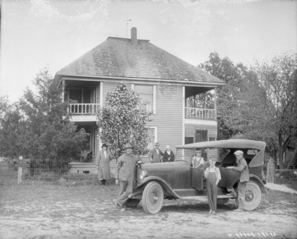 Family posing with car. A man and three children are posing with the car, and behind them three people, a man and two women, are leaning against a wire fence. In the background is a large, two-story house with porches.