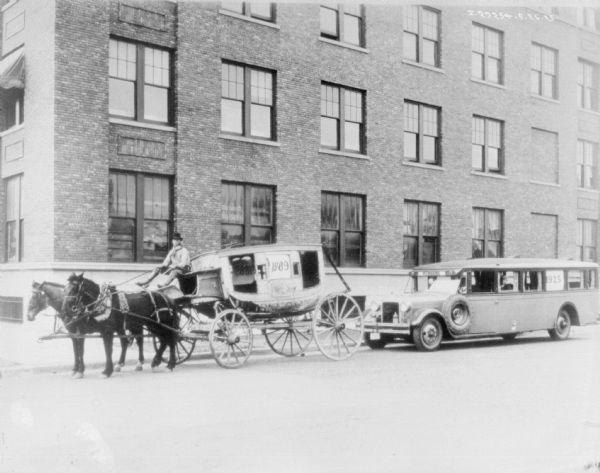 View across street towards a man driving a horse-drawn carriage in front of an International Bus. There is a sign in the carriage that reads: "1889," and a sign in the bus that reads: "1925." Behind them is a large brick building.