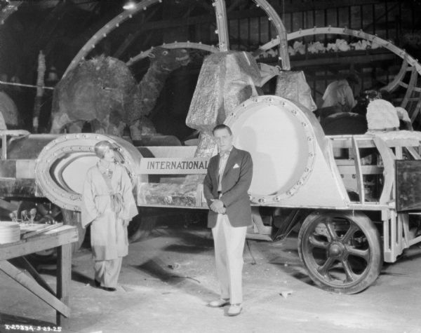 A man wearing a suit and bow tie, and a woman wearing a Kimono-style jacket, and pants and a turban, are standing indoors in front of an International float being built on a trailer. A man is in the background working on the float, which includes a sculpture of a camel, and two sphinxes.
