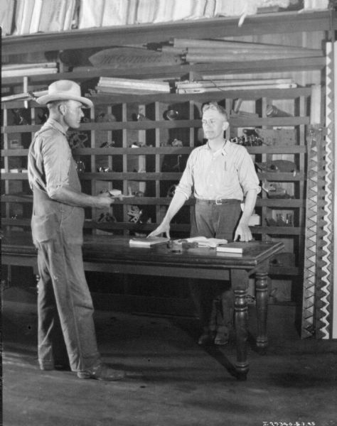 Staged set up showing a salesman, standing behind a table, selling a machine part to a customer. Behind the men are rows of divided shelves with parts in stock.