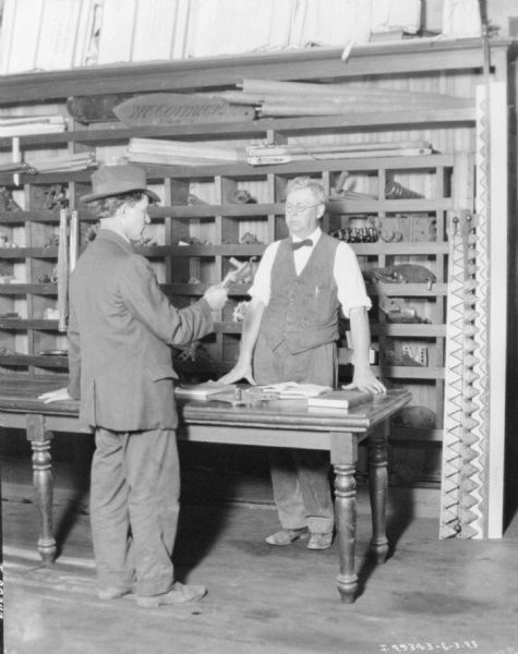 Staged set up showing a salesman, standing behind a table, selling a machine part to a customer. Behind the men are rows of divided shelves with parts in stock.