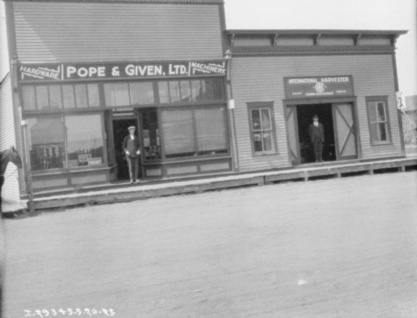 View across road towards a man posing in the open doorway of a storefront with large front windows on the left. The sign above the storefront reads: "Pope & Given, Ltd." There is a horse standing at the corner of the building on the far left. Another man is posing in the large open doorway of the building next door, with a sign above reading: "International Harvester Farm Machines, Sales, Service."
