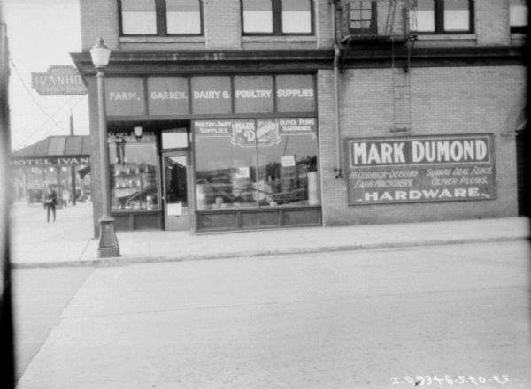 View from across street towards a small corner grocery store. Signs in the window read: "Farm, Garden, Dairy & Poultry Supplies," and "Mark Dumond, McCormick-Deering, Farm Machinery, Square Deal Fence, Oliver Plows, Hardware." 