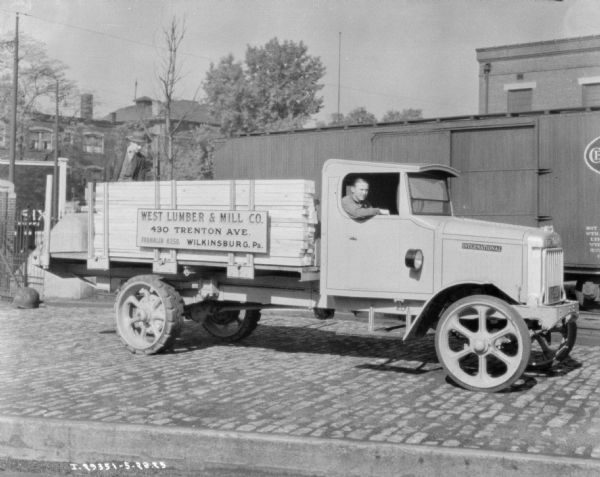 Left side view of truck, with a man posing in the open window of the passenger side door. Another man is standing at the back of the truck bed behind the stacked lumber. There is a railroad car in the background. The sign on the truck bed reads: "West Lumber & Mill Co., 430 Trenton Ave., Wilkinsburg, Pa."