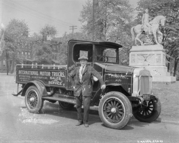 Man posing in front of the passenger door of an International Motor Truck which is parked on a street. In the background on the right is a monument of a man riding a horse.