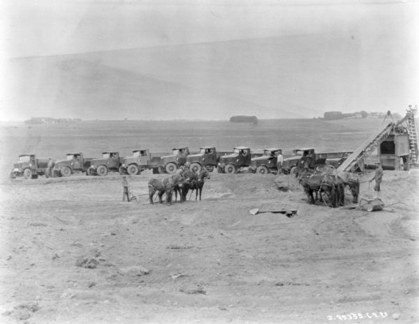 Elevated view of a quarry. Two men in the center are standing behind teams of four horses pulling equipment. Behind them are men posing in trucks parked at an angle in a row. On the right is a piece of conveying machinery.