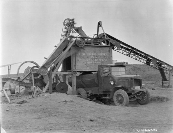 Truck outdoors under a building with conveying machinery. A sign painted on the machinery reads: "5 YD LOADING HOPPER MFD. BY RUSSEL GRADER MFG. CO."