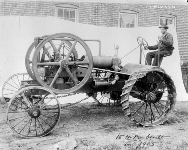 Left side profile view of a man sitting on a 15 HP tractor. There is a backdrop against a brick factory wall in the background. Written at the bottom: "15 H.P. built in 1905."