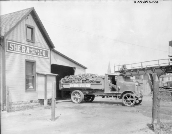A delivery truck with a pickup bed piled with material is sitting in the open doorway of the Shera Coal & Oil Co. building. A man is sitting in the open doorway of the passenger side of the truck. Above and to the right is a section of conveying machinery. In the far background is a church steeple.