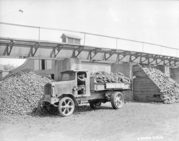 A man is sitting in the open doorway of the driver's seat of a truck at the Shera Coal & Oil Co. Near the truck is a pile of material, and above and behind is conveying machinery. There is a large wood building in the background.