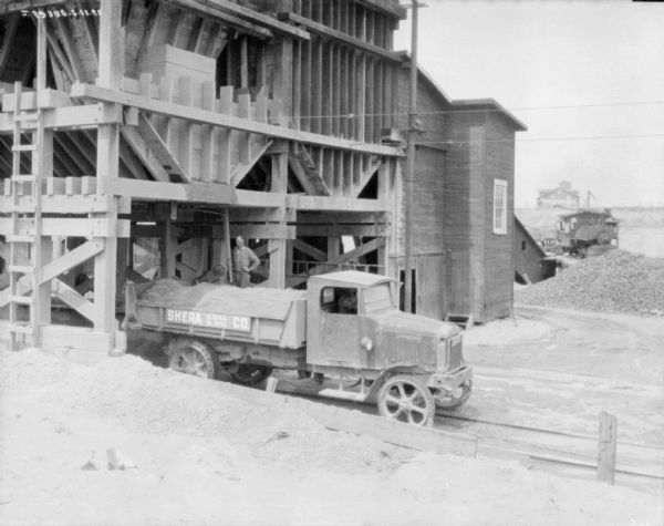 View looking down at a man standing under a hopper near the side of a truck for the Shera Coal & Oil Co. A man is sitting in the driver's seat of the truck.