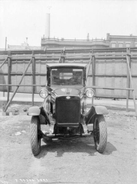 View from front of a truck parked in front of a tall, sturdily supported wood fence. There are three-story brick buildings and a tall smokestack in the background.
