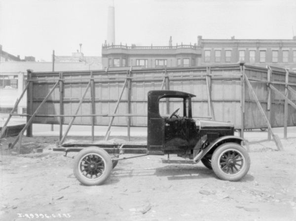 Right side view of a truck parked in front of a tall, sturdily supported wood fence. There are three-story brick buildings and a tall smokestack in the background.