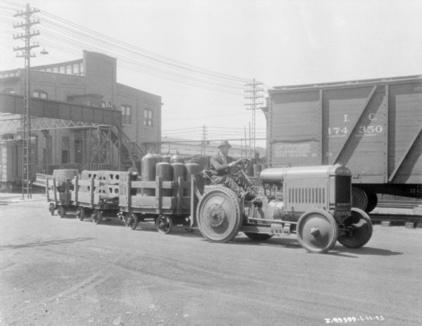 A man is driving an industrial tractor pulling a train of carts. The carts are loaded with parts, tanks, and barrels. In the background are factory buildings, and on the right is a railroad car.