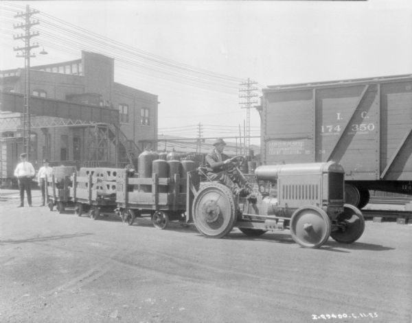 A man is driving an industrial tractor pulling a train of carts. The carts are loaded with parts, tanks, and barrels. In the background are factory buildings, and on the right is a railroad car. Two men are standing on the left near one of the carts.