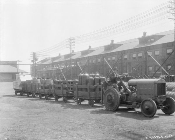 A man is driving an industrial tractor pulling a train of carts. The carts are loaded with parts, tanks, and barrels. In the background are factory buildings.