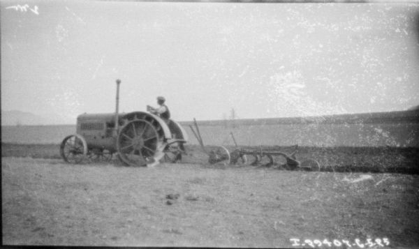 Left side profile view of a man driving a 10-20 tractor pulling a plow in a field.