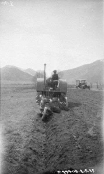 Rear view of a man using a 10-20 tractor to pull a plow in a field. Hills are in the background. There is an automobile parked on the right in the background.