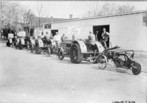 Three-quarter view from left rear of four men parading farm implements in a street next to a long building. There are a number of signs that read, in part: "This Tractor ___ by F.C. Putney..." Men are standing in front of the building watching.