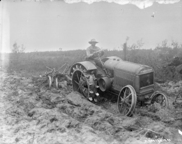Three-quarter view from front right of a man using a tractor for pulling a plow to break up a field.